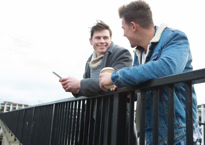 Two young men talking in the city. One is holding a smartphone, the other is holding a disposable coffee cup.